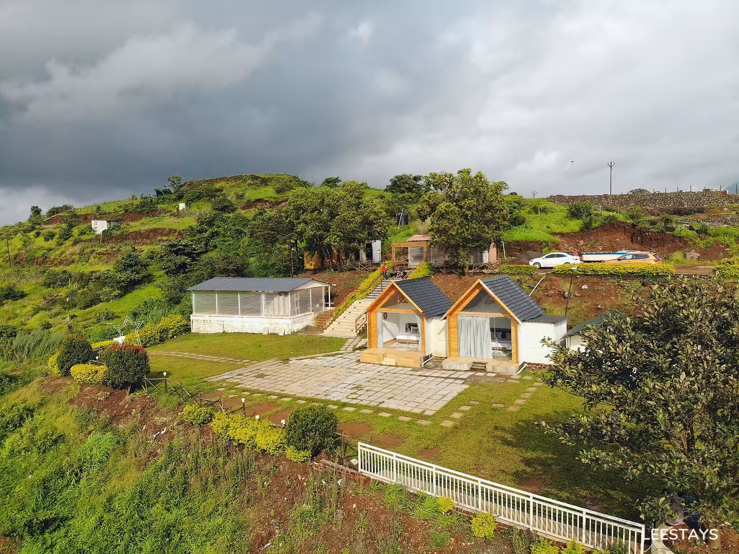 Two small cabins with pitched roofs and open fronts on a paved area, with a screened enclosure and a hillside behind.