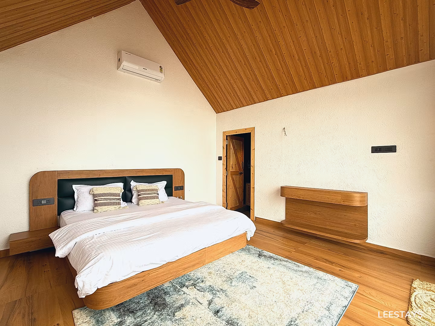 Wooden bed with white linens and decorative pillows, facing a textured wall with an air conditioner and a wooden shelf.