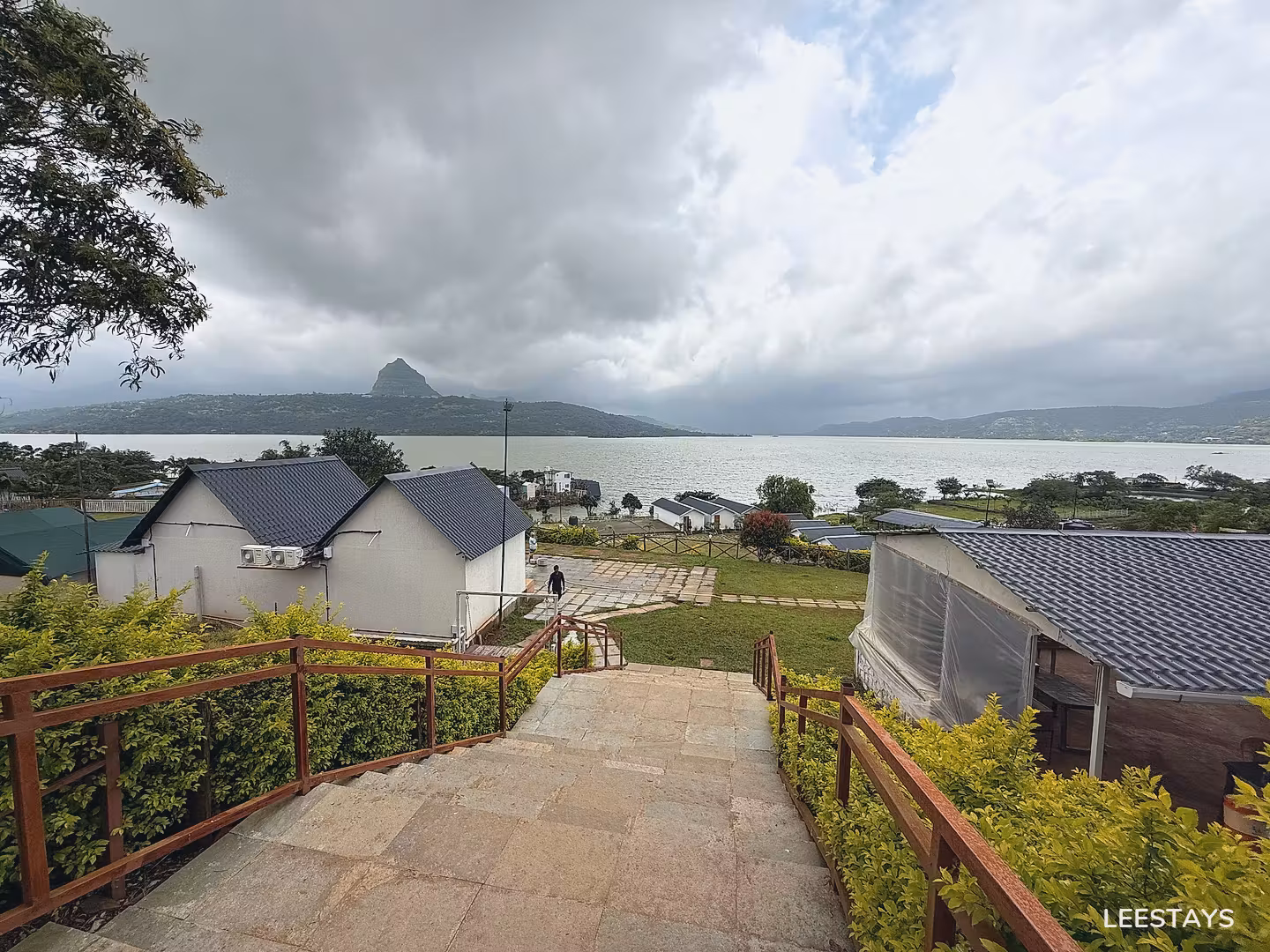 Stone steps with metal railings lead down to white cottages with dark roofs overlooking a large lake and distant hills.