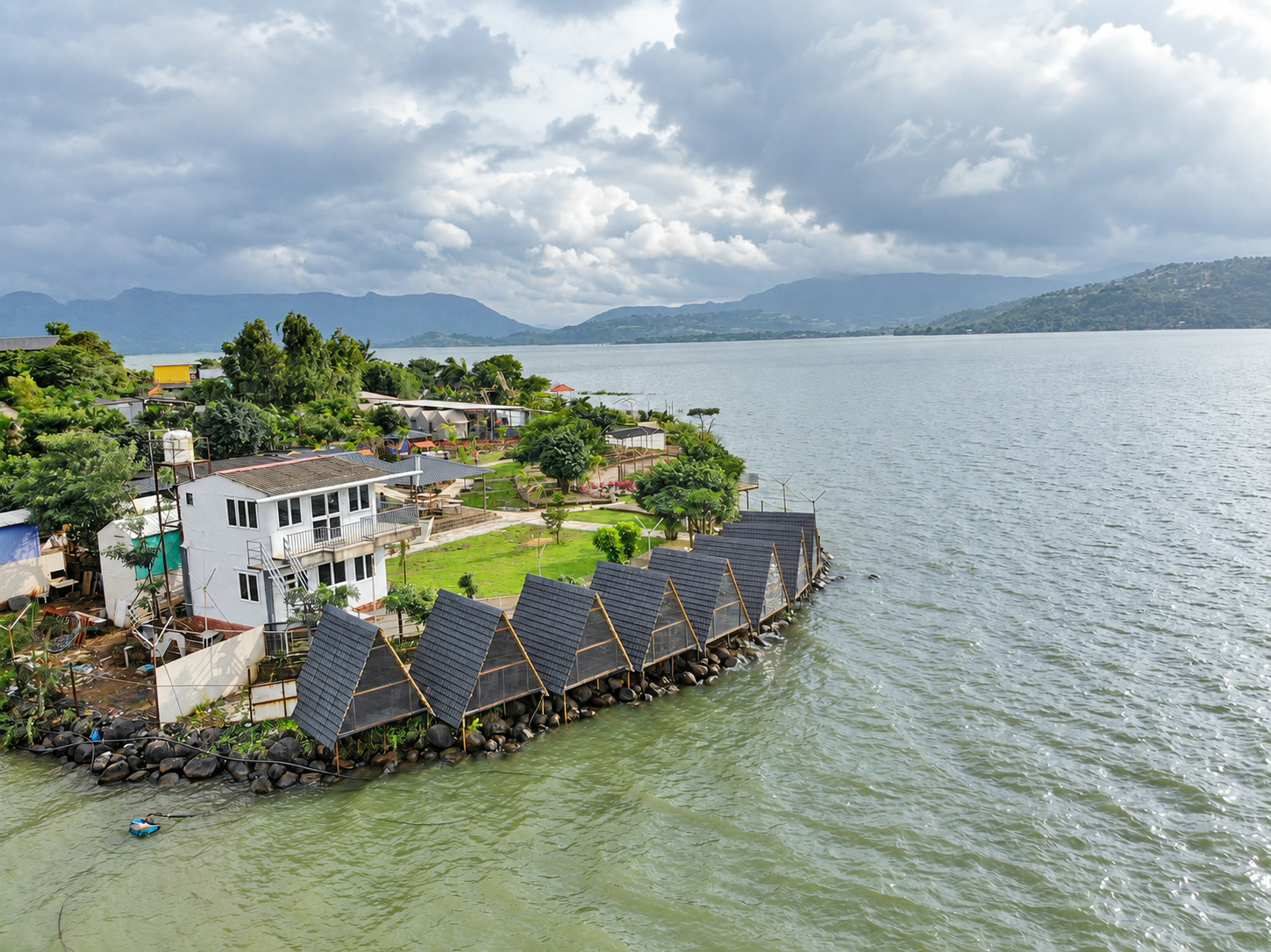 Wide drone view of NightVibe triangle cabins, gardens and Pawna Lake.