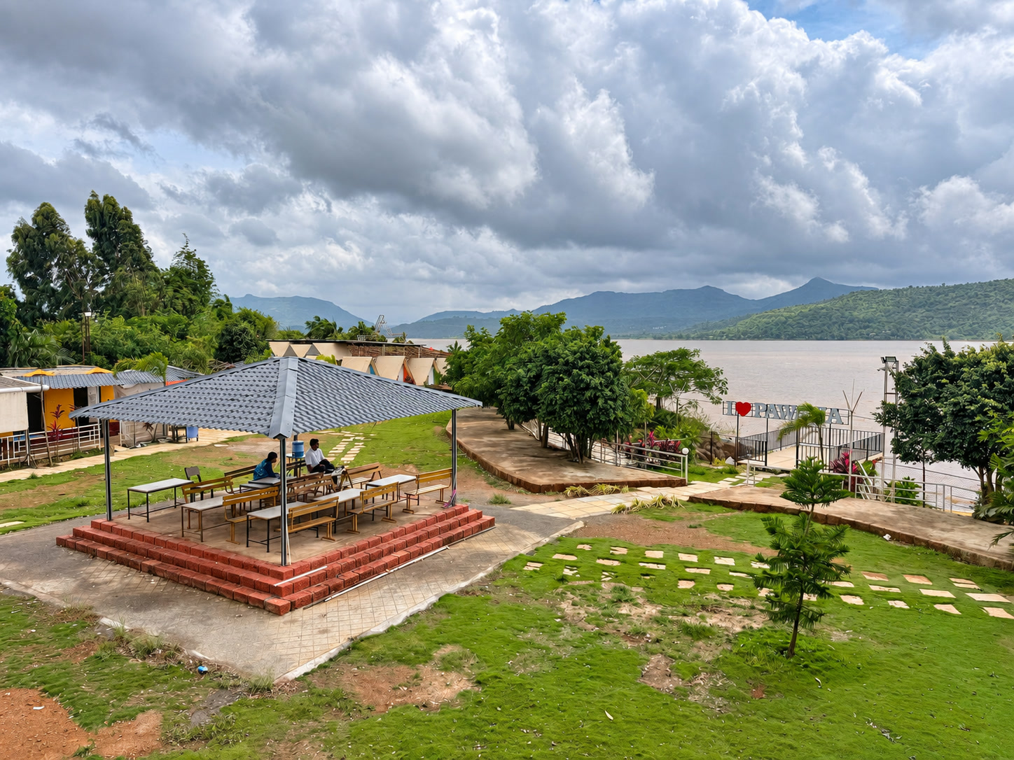 A covered outdoor pavilion with tables and benches sits on a raised brick platform amidst grassy grounds with stepping stones. The property overlooks a large body of water and distant hills under an overcast sky, with small cabins and triangular structures visible in the background.