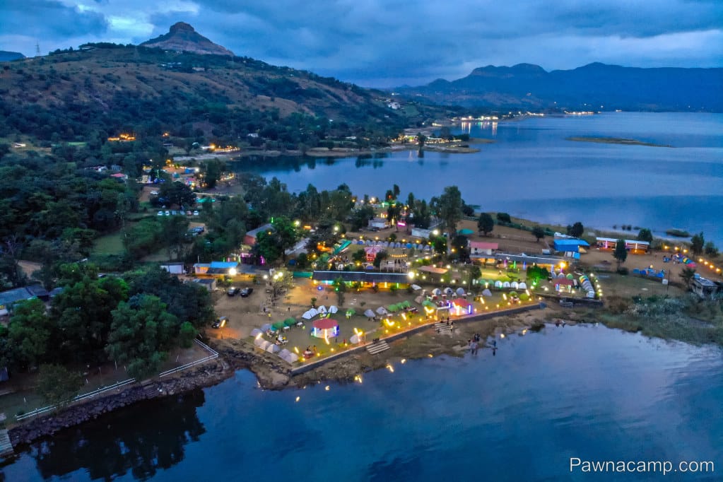 Aerial view of a lakeside camping area at Pawna Lake, featuring tents and illuminated pathways along the shore.