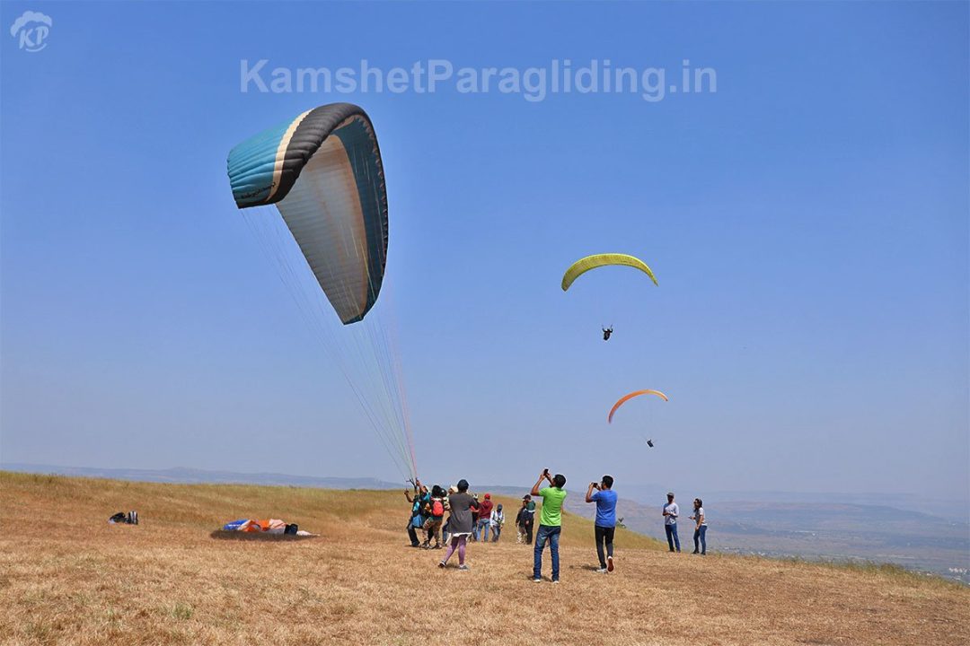 Paragliders glide over Kamshet while pilots and spectators gather on the hillside, with one wing descending toward landing below.