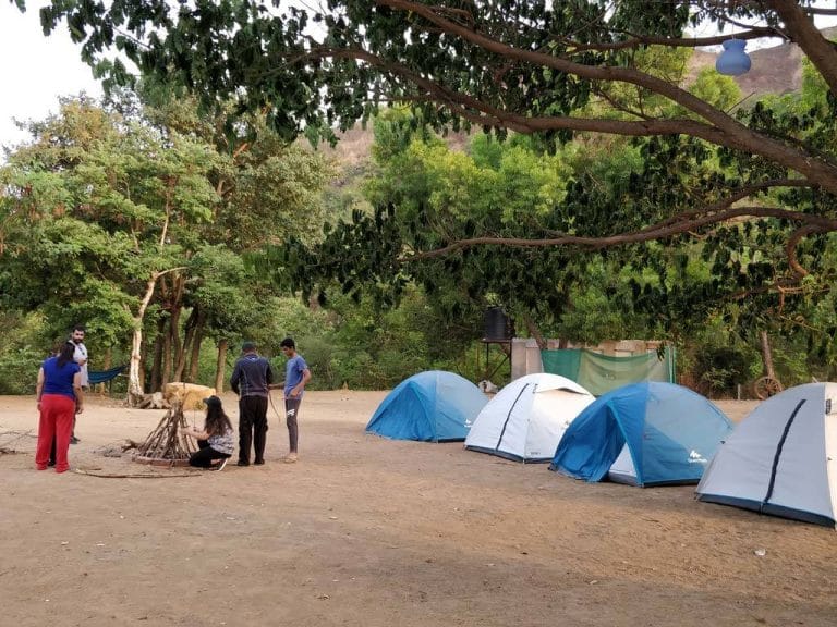 Several blue camping tents arranged in a natural setting with people gathered around a fire pit.