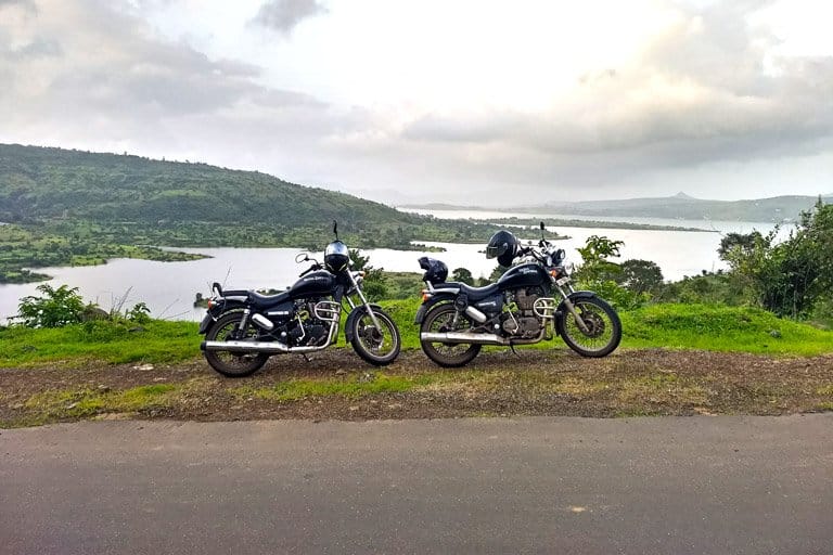 Two black motorcycles parked on a roadside with a view of Pawna Lake and surrounding hills in the background.