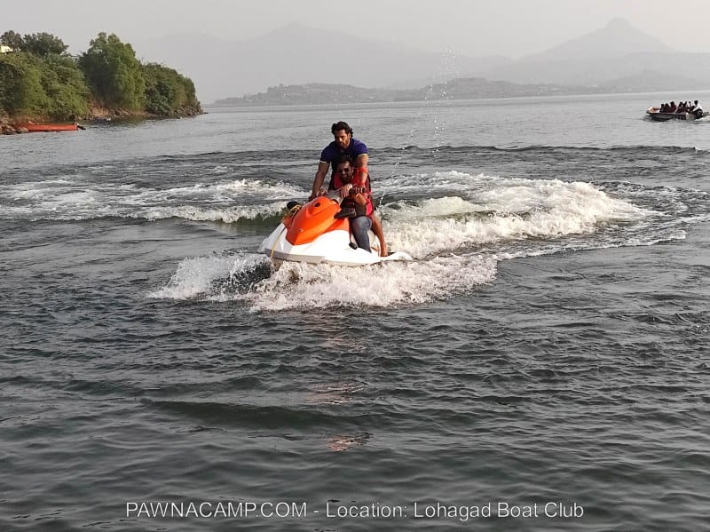 Two individuals riding a jet ski on the water at Pawna Lake, creating waves behind them.