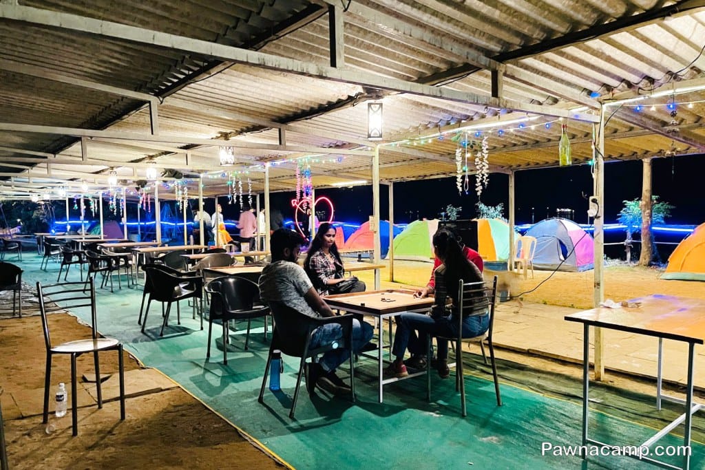 Outdoor seating area with tables and chairs, illuminated by decorative lights, and tents in the background.