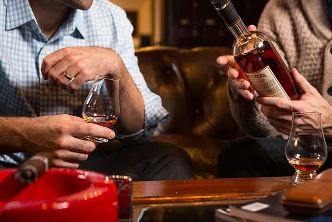 Two people holding glasses of whiskey in a cozy setting, suitable for a relaxing stay near Pawna Lake.