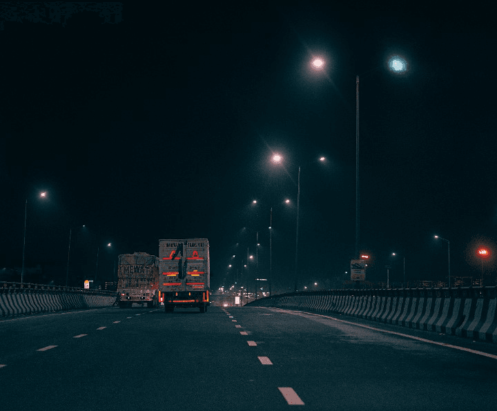 A dimly lit highway at night with trucks traveling, suggesting a journey to Pawna Lake for a stay amidst tranquil surroundings.