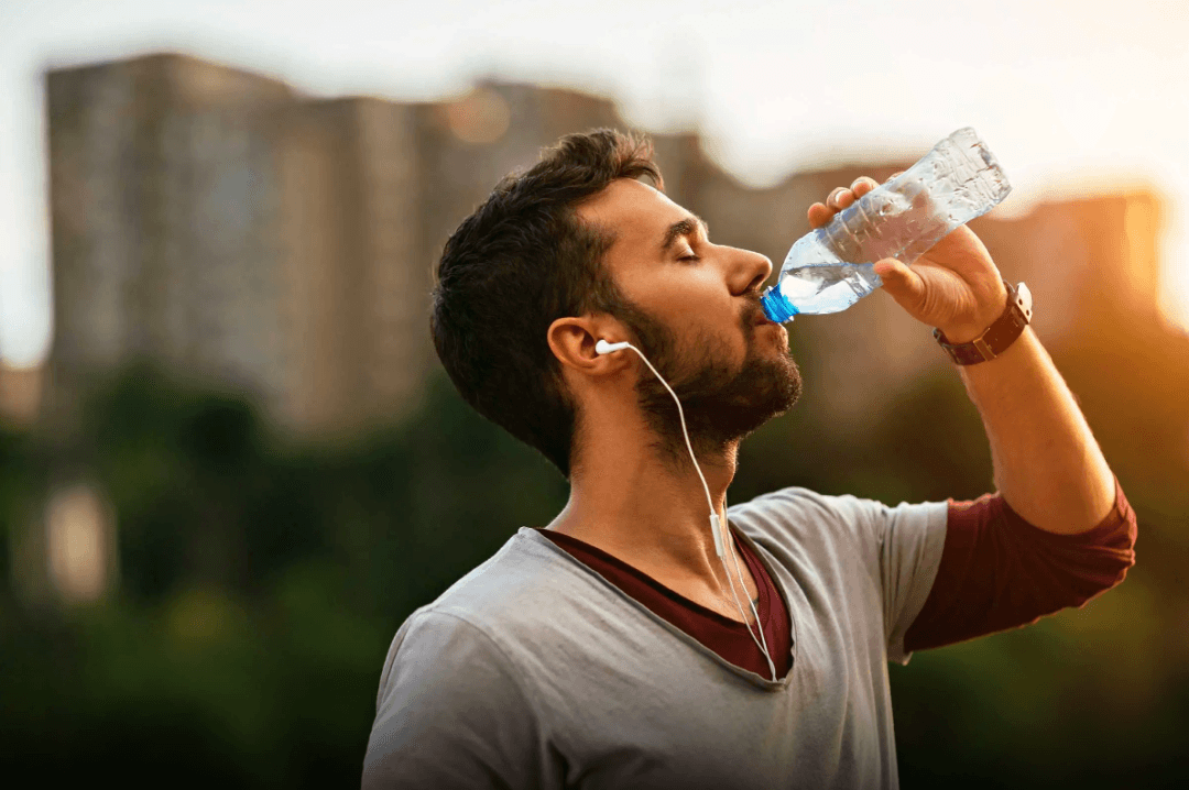 A person drinking from a water bottle with earphones on, set against a blurred backdrop reminiscent of Pawna Lake stay scenery.