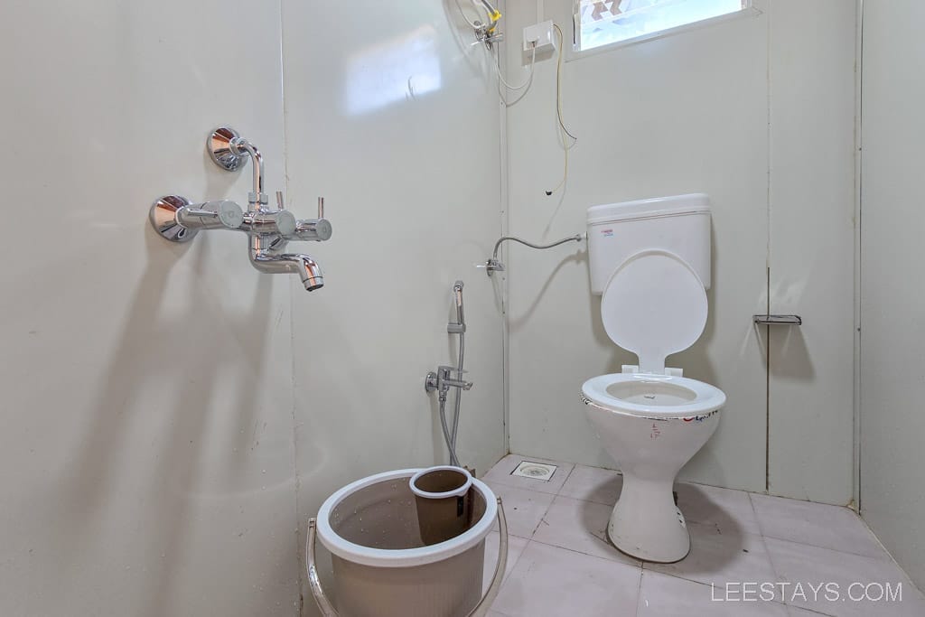 Simple bathroom setup in a budget stay near Pawna Lake, featuring a toilet, shower faucet, and bucket with mug.